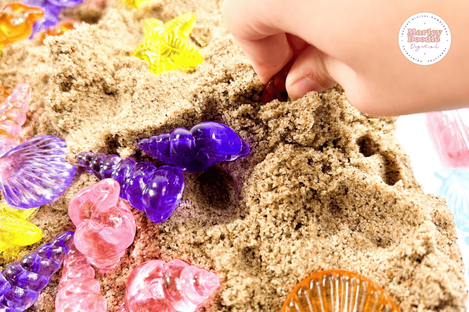 Child digging through sand to find colorful seashell toys in a sensory bin.