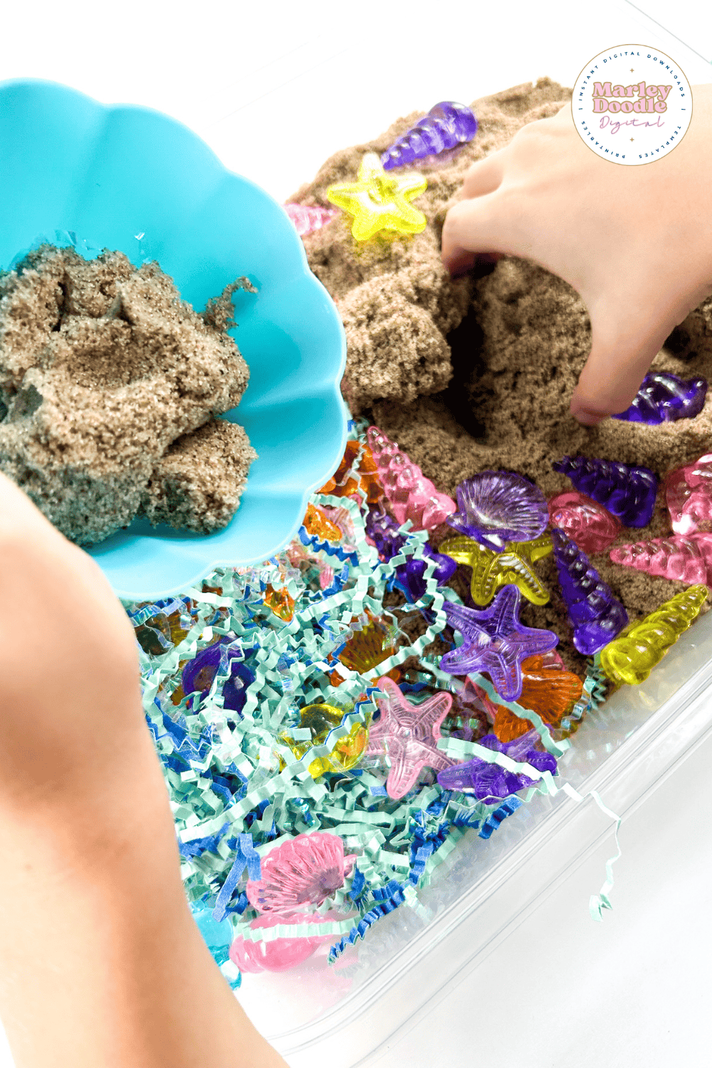 Child scooping kinetic sand into a blue bowl in a seashell sensory bin.